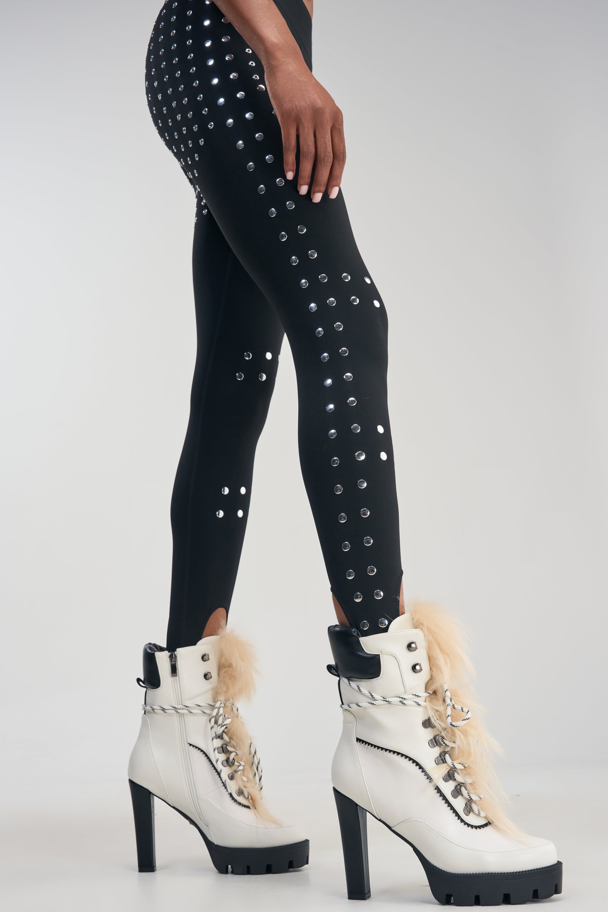 A person wearing black leggings with silver studs and the AKIRA GOLD SNUG WHITE FUR BLOCK HEEL COMBAT BOOTIE stands against a plain light background, showcasing the cozy elegance of this stylish white lace-up bootie with fur trim and a modern chunky lug sole.