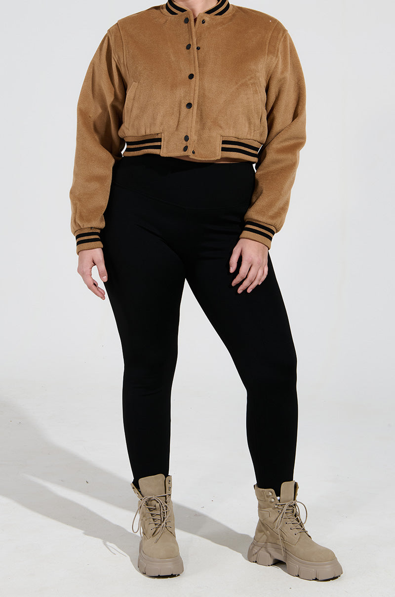A person is wearing a tan bomber jacket with black trim, paired with SNUGGLE FLEX Fleece Leggings in Black and tan combat boots, standing against a neutral background.