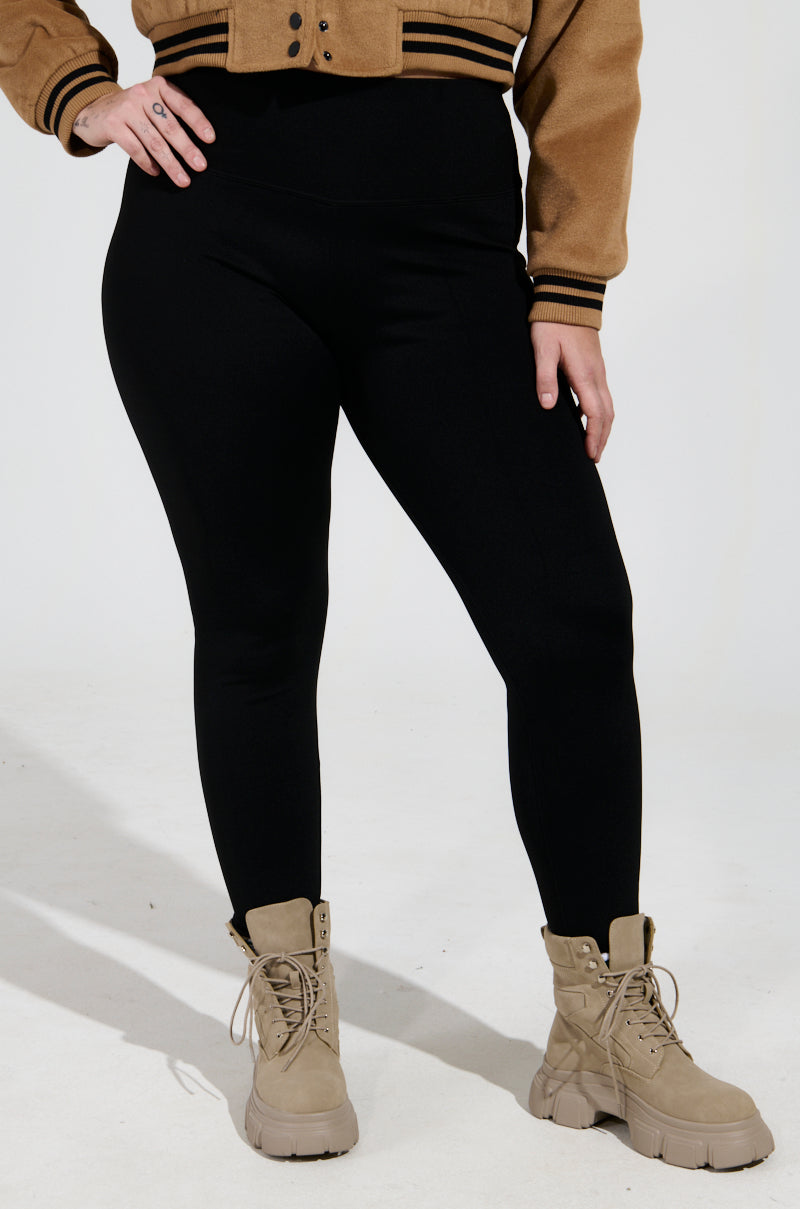 A person wearing the Snuggle Flex Fleece Legging Extended Size in black, paired with a brown jacket and beige lace-up boots, stands against a plain background with hands at their sides, highlighting a casual and stylish ensemble.