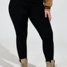 A person wearing the Snuggle Flex Fleece Legging Extended Size in black, paired with a brown jacket and beige lace-up boots, stands against a plain background with hands at their sides, highlighting a casual and stylish ensemble.