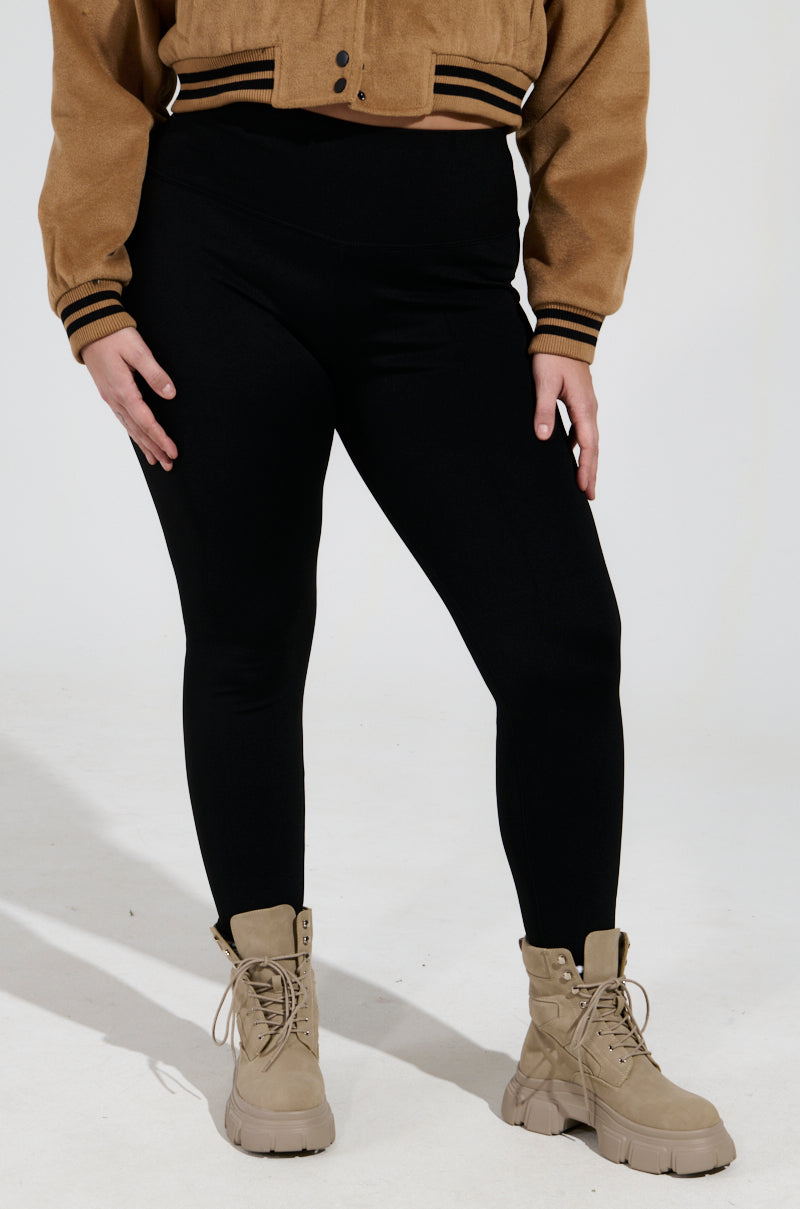 A person wearing SNUGGLE FLEX FLEECE LEGGING EXTENDED SIZE IN BLACK, a tan jacket, and beige combat boots. The image is cropped to show only from the shoulders down against a plain white background.
