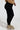 A person wearing the Snuggle Flex Fleece Legging Extended Size in black, a tan jacket, and beige chunky boots stands against a plain light background. The person is positioned with their right side facing the camera.
