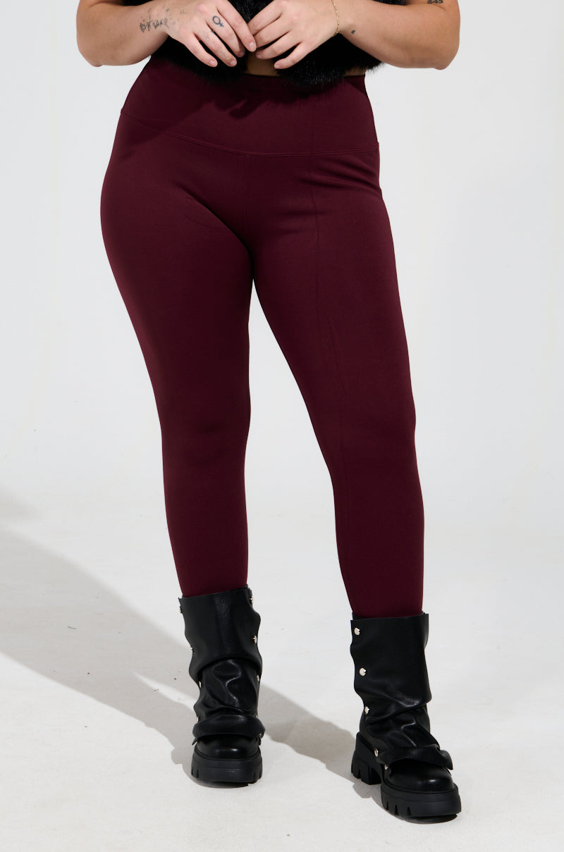 A person is showcasing the SNUGGLE FLEX FLEECE LEGGING EXTENDED SIZE in burgundy, paired with black leather boots adorned with metal studs, against a plain white backdrop. Only their lower torso and legs are visible.