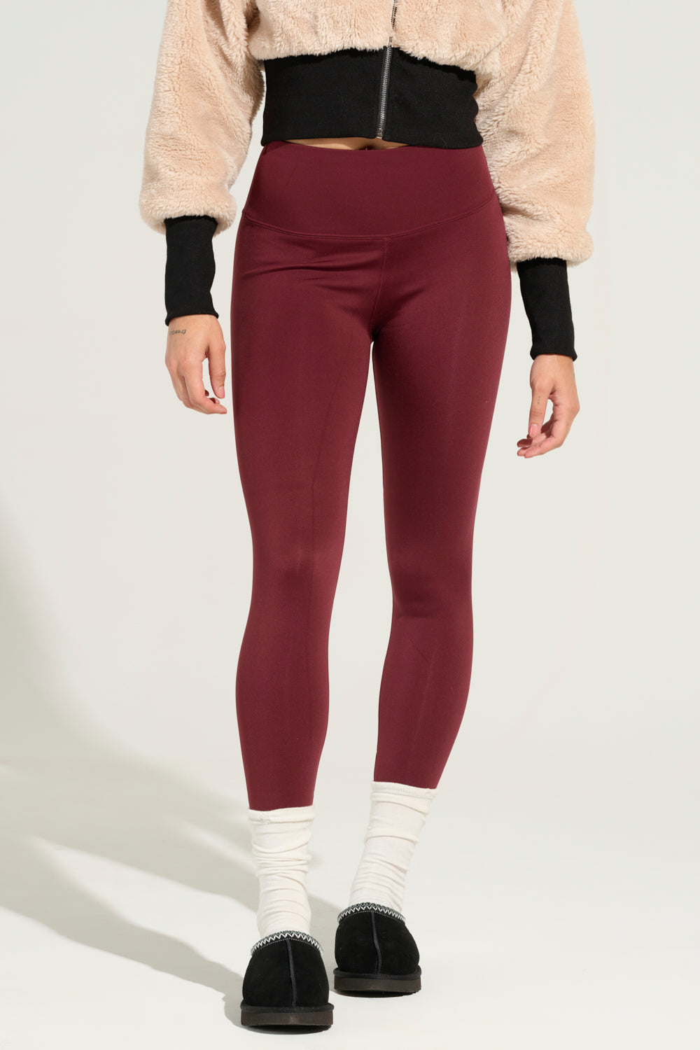A person wearing a beige fuzzy jacket, SNUGGLE FLEX FLEECE LEGGING IN RED, white socks, and black slippers stands against a light background. They are facing the camera with their hands relaxed at their sides. 1
