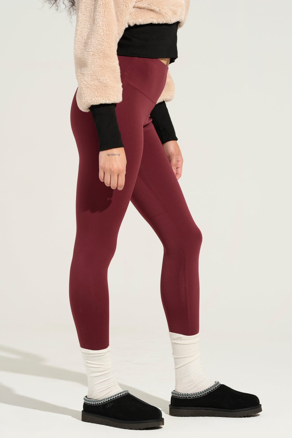 A person wearing Snuggle Flex Fleece Legging in Red, a beige fuzzy sweater, black top underneath, white socks, and black slip-on shoes stands against a plain background. Only the lower half of the body is visible. 2