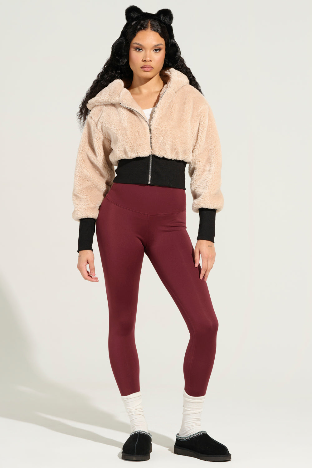 A woman poses in a beige fuzzy jacket with a zipper, wearing SNUGGLE FLEX FLEECE LEGGINGS IN RED, white socks, and black slip-on shoes. Her long, wavy hair is adorned with black cat ear accessories against a plain light background. 4