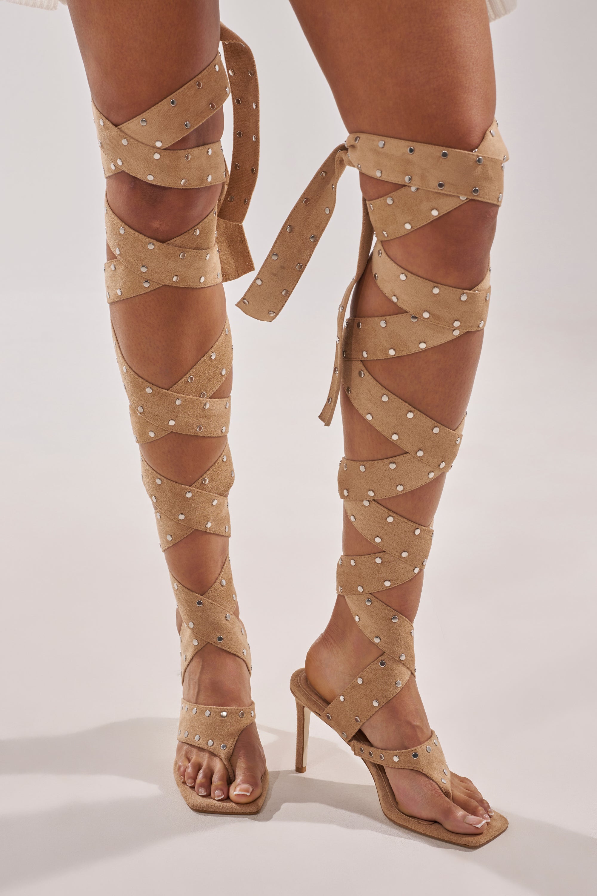 A person wears the AZALEA WANG SODALITE NUDE FAUX SUEDE LACE UP SANDAL, featuring nude faux suede, lace-up design with silver studs, a square toe heel, and straps wrapping up to just below the knees. 0