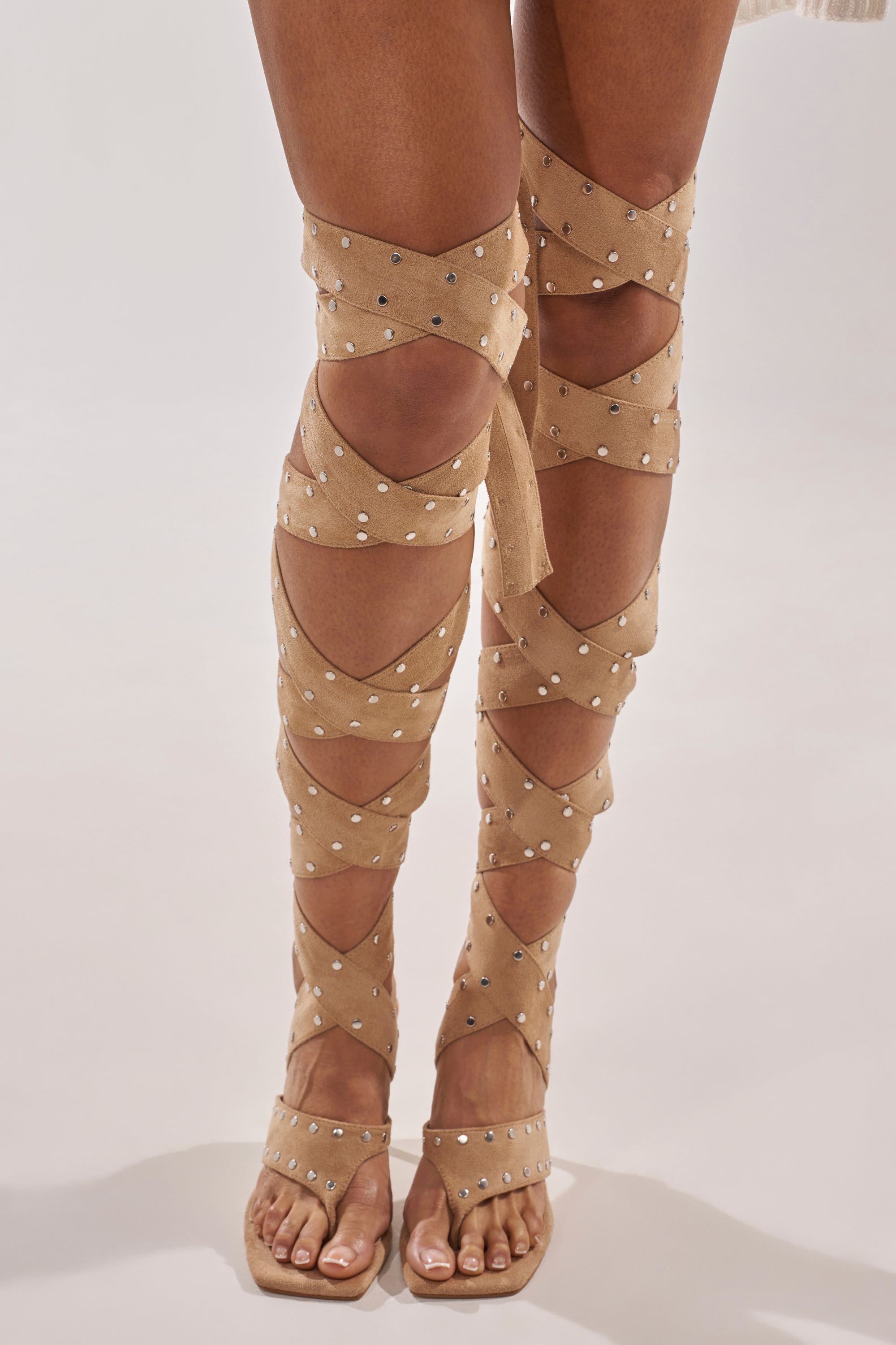 Someone wears the AZALEA WANG SODALITE NUDE FAUX SUEDE LACE UP SANDAL, featuring crisscross faux suede straps with small silver studs wrapping up to the knees. Toes are visible against a plain, light-colored background. 2