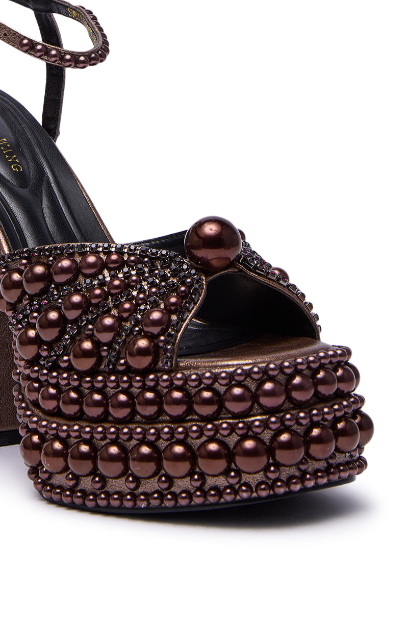 The AZALEA WANG SOMETHING BRONZE EMBELLISHED PLATFORM SANDAL features a close-up of its embellished heel with shiny bronze pearls and beads, intricate patterns, a peep toe, and a sleek black ankle strap.