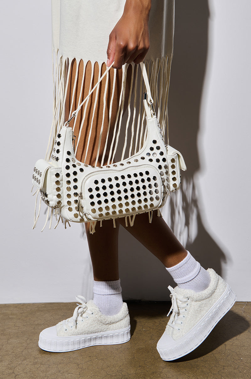A person in a white fringe dress holds a white handbag with circular cutouts. Their AZALEA WANG SOSI WHITE SNEAKER, featuring a flatform sole and paired with white socks, rests on a brown textured floor, with soft lighting highlighting the outfit's clean lines and subtle details.