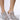 Close-up of a person wearing the AZALEA WANG SOULMATE SILVER EMBELLISHED PUMP WITH RIBBON DETAIL—sparkly high-heeled mules adorned with rhinestone detailing, iridescent sequins, pink laces, and jewels. The shoes have a pointed toe design, and the person's feet are positioned on a smooth, light-colored surface.
