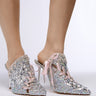 Close-up of a person wearing the AZALEA WANG SOULMATE SILVER EMBELLISHED PUMP WITH RIBBON DETAIL—sparkly high-heeled mules adorned with rhinestone detailing, iridescent sequins, pink laces, and jewels. The shoes have a pointed toe design, and the person's feet are positioned on a smooth, light-colored surface.