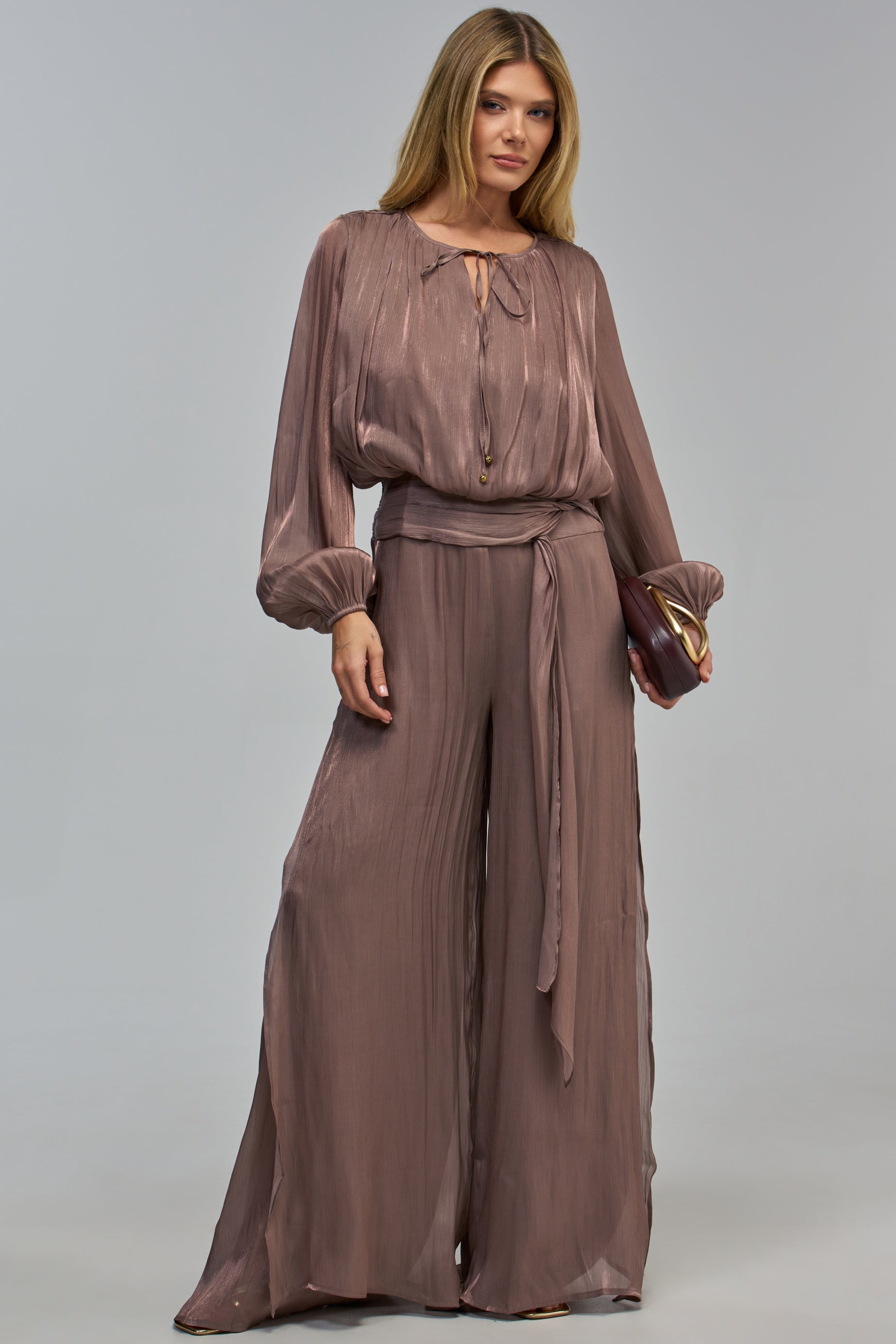 A woman wears a FAVORITE DAUGHTER CHIFFON JUMPSUIT IN MOCHA, an elegant, long-sleeved mocha chiffon jumpsuit with a pleated finish, a tied waist, and a dark-colored clutch.