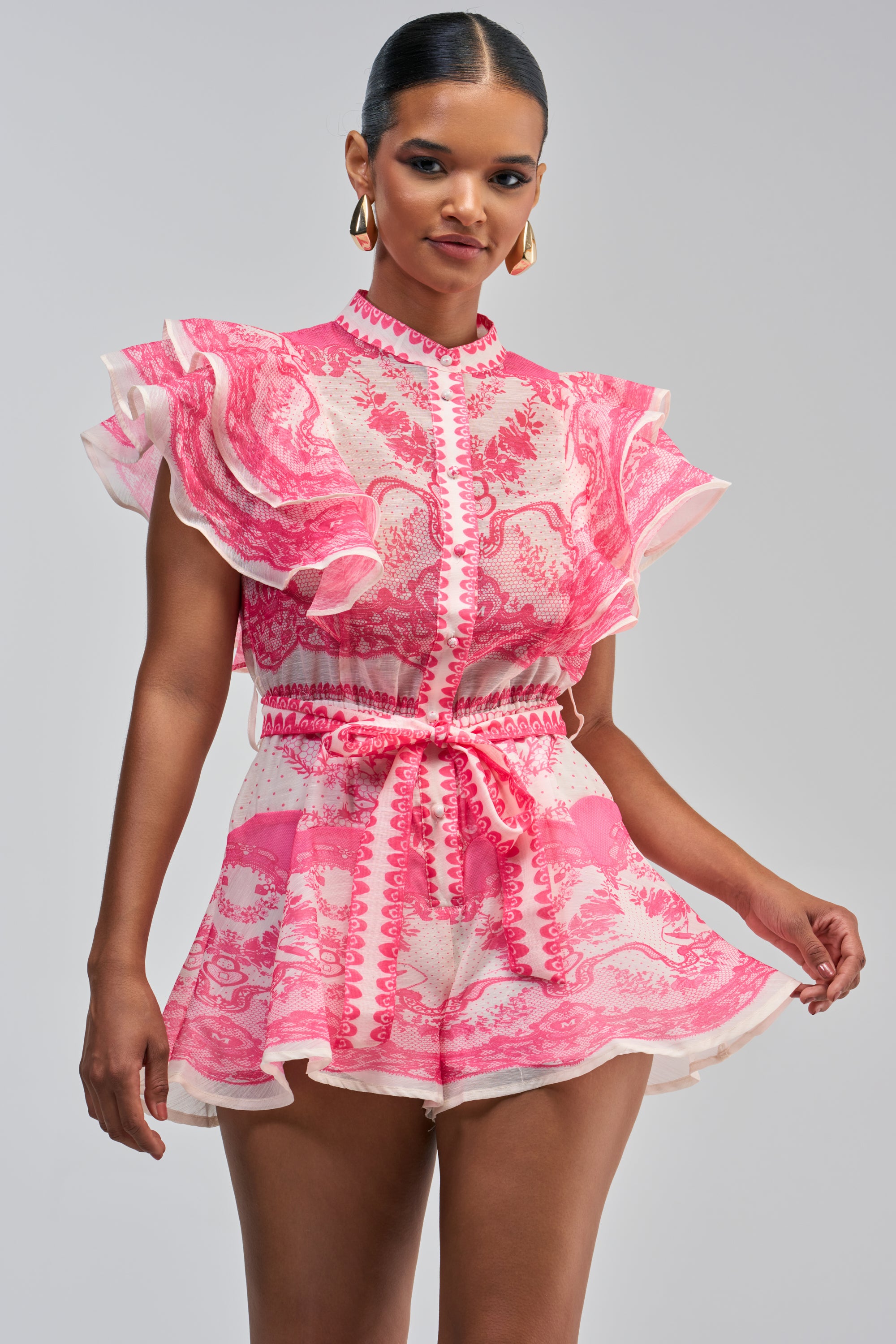 A woman models the PRETTY IN PINK PATTERNED ROMPER—a short, pink and white romper featuring ruffle detailing on the sleeves and a tied waist—posing against a light gray background. Her slicked-back hair and gold earrings complete this chic brunch-ready look.