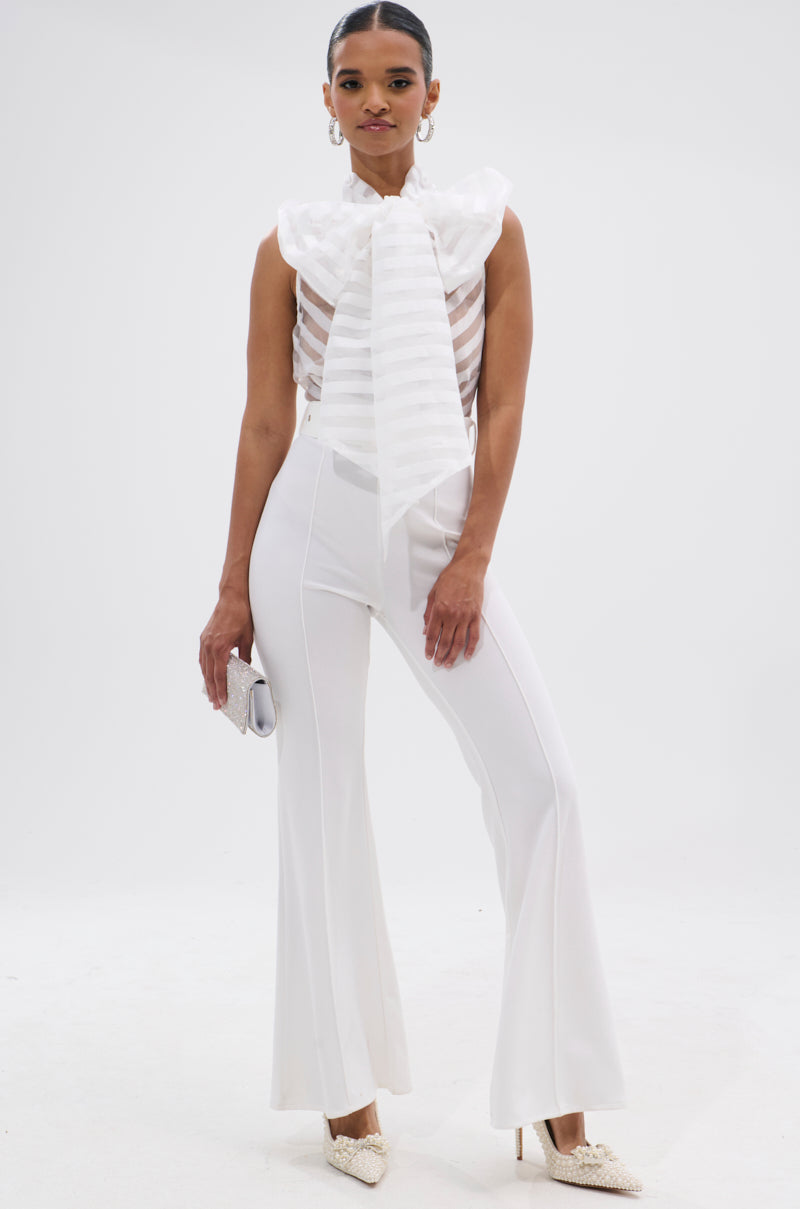 A woman poses against a white background in the GIRLY POP JUMPSUIT IN WHITE, featuring a sleeveless design with a statement bow and wide legs, paired with pointed white heels and holding a small clutch.