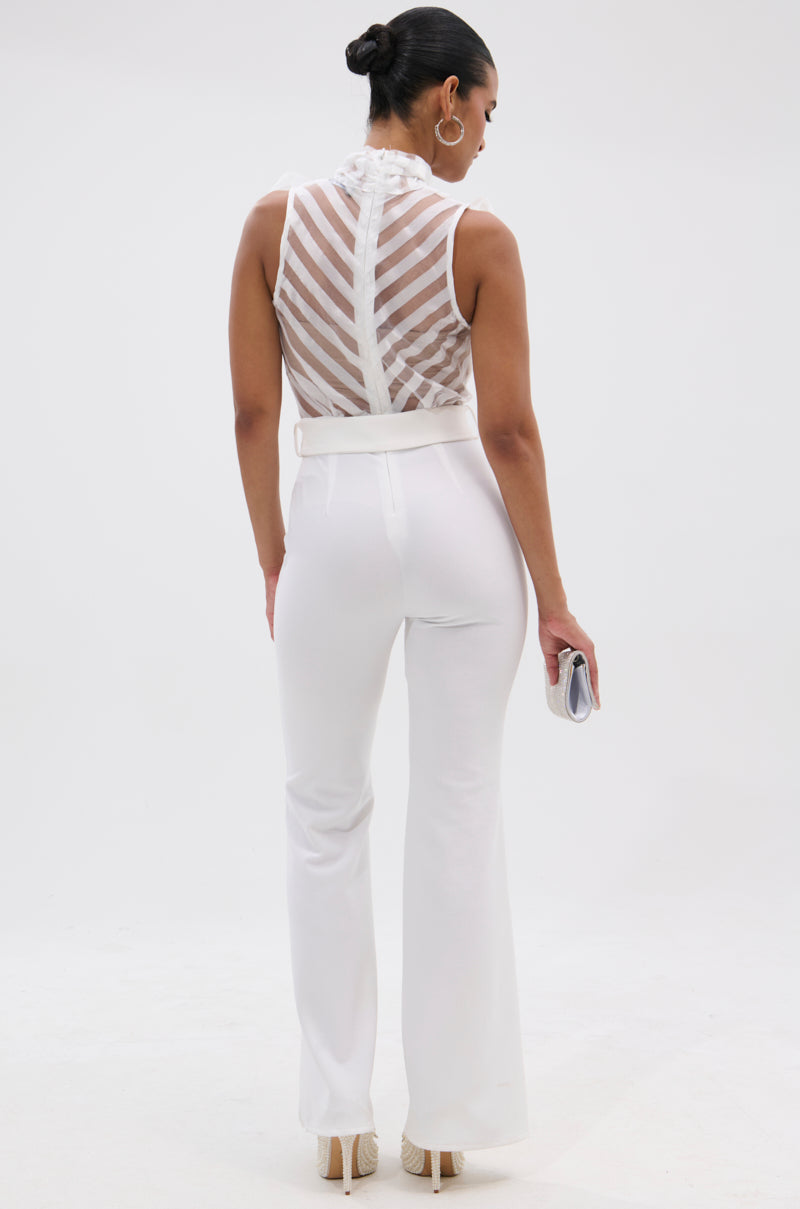 A woman faces away, wearing the GIRLY POP JUMPSUIT IN WHITE. She holds a small clutch, wears hoop earrings and heels, with her hair styled in a sleek bun.