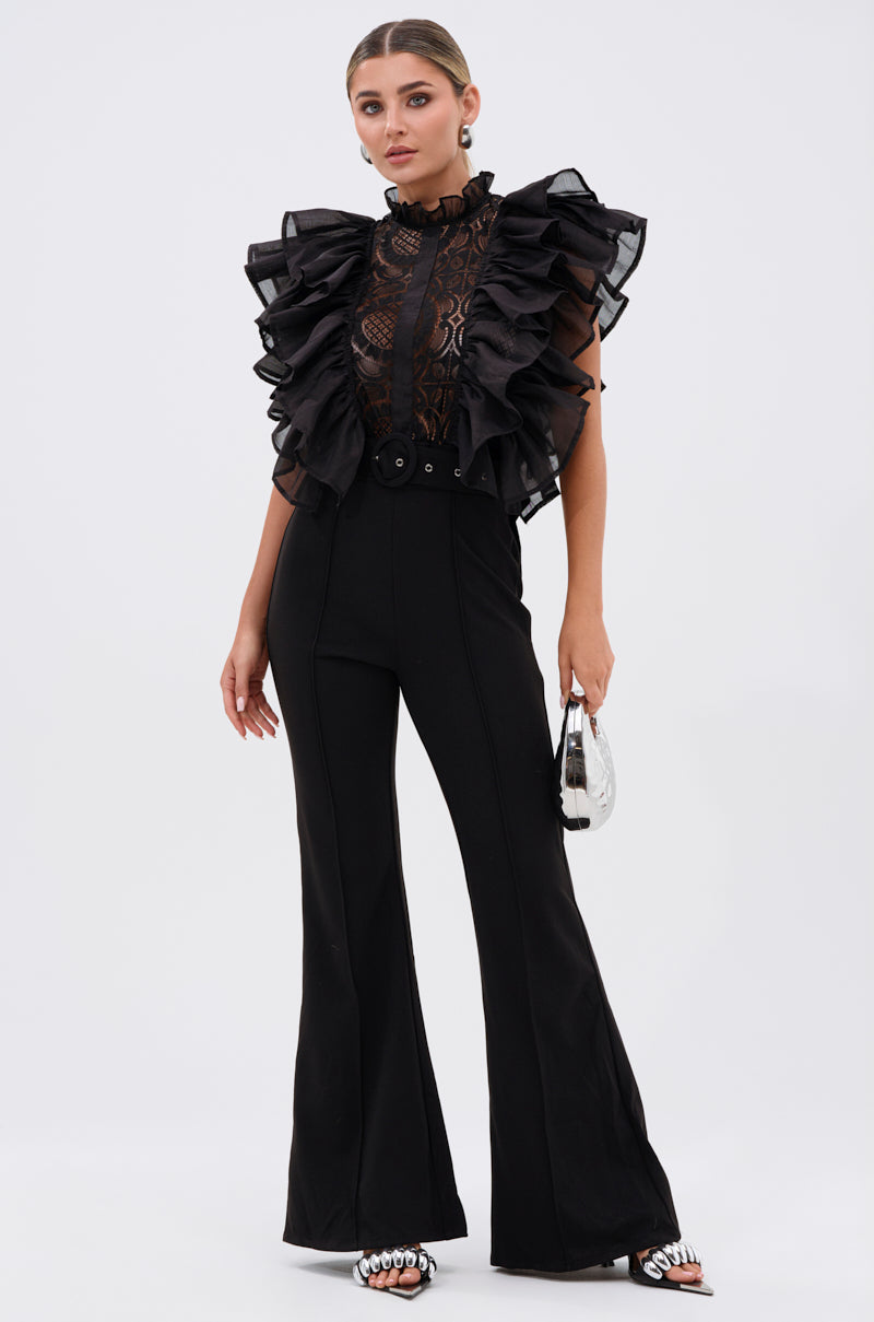 Woman stands against a plain white background wearing the WALK THAT WALK RUFFLE JUMPSUIT IN BLACK, styled with a small silver bag and jeweled open-toe heels. Her hair is slicked back for a sleek look.