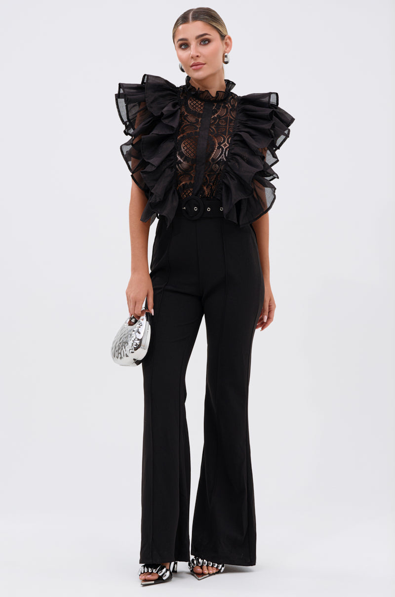 A woman stands against a plain background wearing the WALK THAT WALK RUFFLE JUMPSUIT IN BLACK, paired with a silver handbag and zebra-patterned heels.
