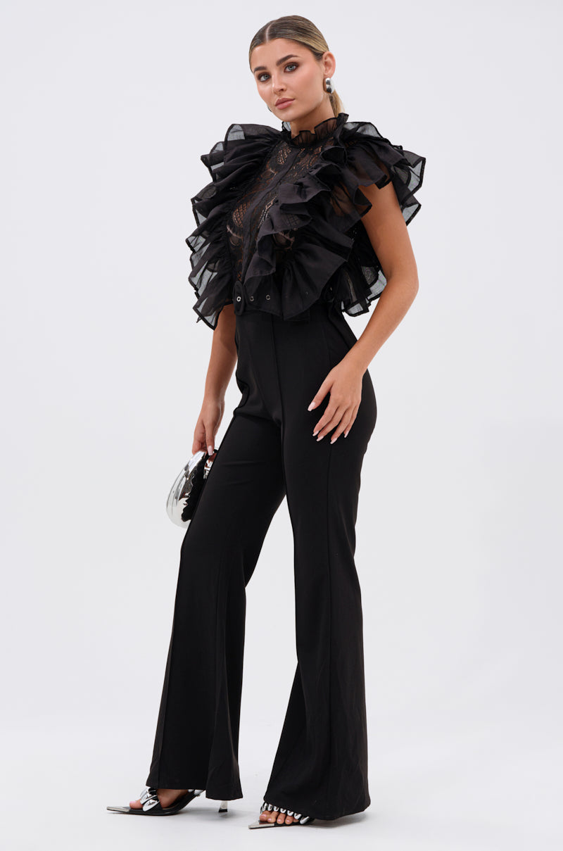 A woman stands against a white background in the WALK THAT WALK RUFFLE JUMPSUIT IN BLACK with high-waisted pants, open-toe heels, and a small silver purse. Her hair is styled back for a polished look.
