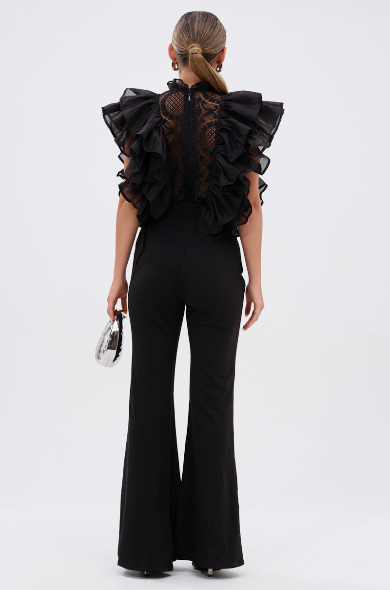 A woman faces away in the WALK THAT WALK RUFFLE JUMPSUIT IN BLACK, featuring lace embroidery and flared sleeves. Her hair is styled in a low ponytail, and she holds a small silver clutch against a plain white background.
