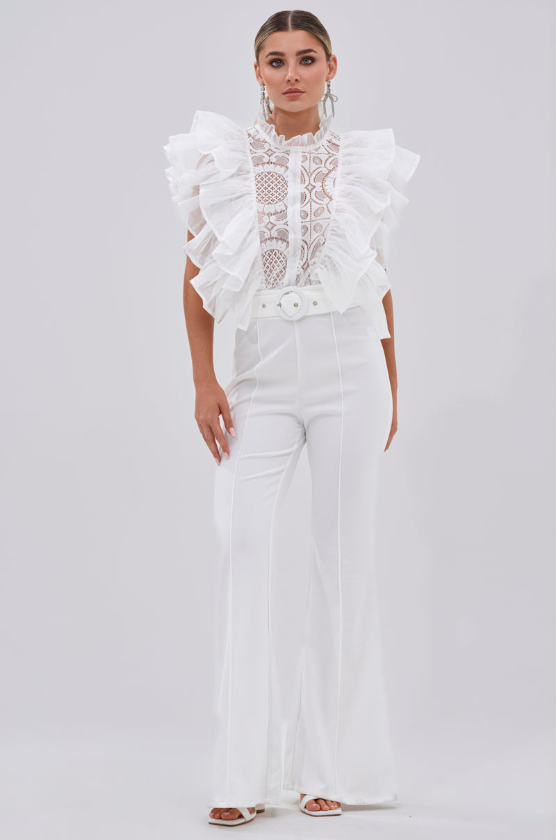 A woman poses against a plain background wearing the WALK THAT WALK RUFFLE JUMPSUIT IN WHITE, featuring lace sleeves, a high neckline, flared pants, and a wide belt with a large buckle. She completes the chic look with hoop earrings and slicked-back hair.