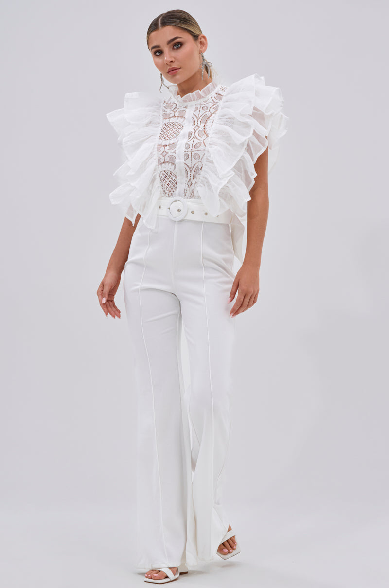A woman poses confidently against a plain background, wearing the WALK THAT WALK RUFFLE JUMPSUIT IN WHITE with a belted waist and open-toed heels. Her hair is styled back, highlighting the chic all-white look.