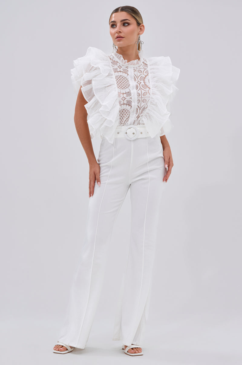 A woman stands confidently in the WALK THAT WALK RUFFLE JUMPSUIT IN WHITE, featuring wide legs, a wide belt, and a sheer ruffled lace top. She pairs it with white sandals, statement earrings, and wears her hair pulled back.