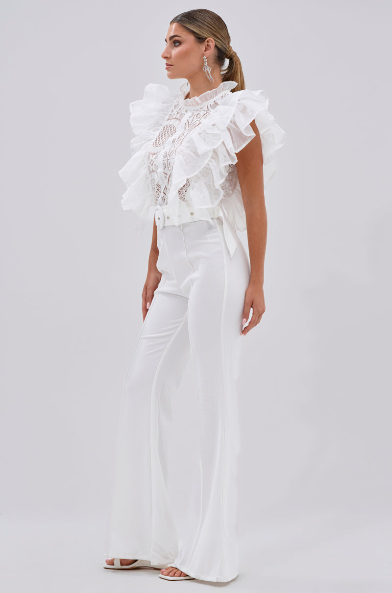 A woman in profile poses against a plain background, wearing the WALK THAT WALK RUFFLE JUMPSUIT IN WHITE with sheer lace panels and wide-leg pants. Her long hair is in a ponytail, paired with white open-toe heels.