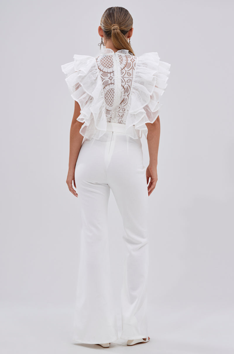 A woman faces away in the WALK THAT WALK RUFFLE JUMPSUIT IN WHITE, featuring chic lace embroidery, sheer ruffled sleeves, and wide legs. Her hair is in a sleek ponytail against a plain light background.