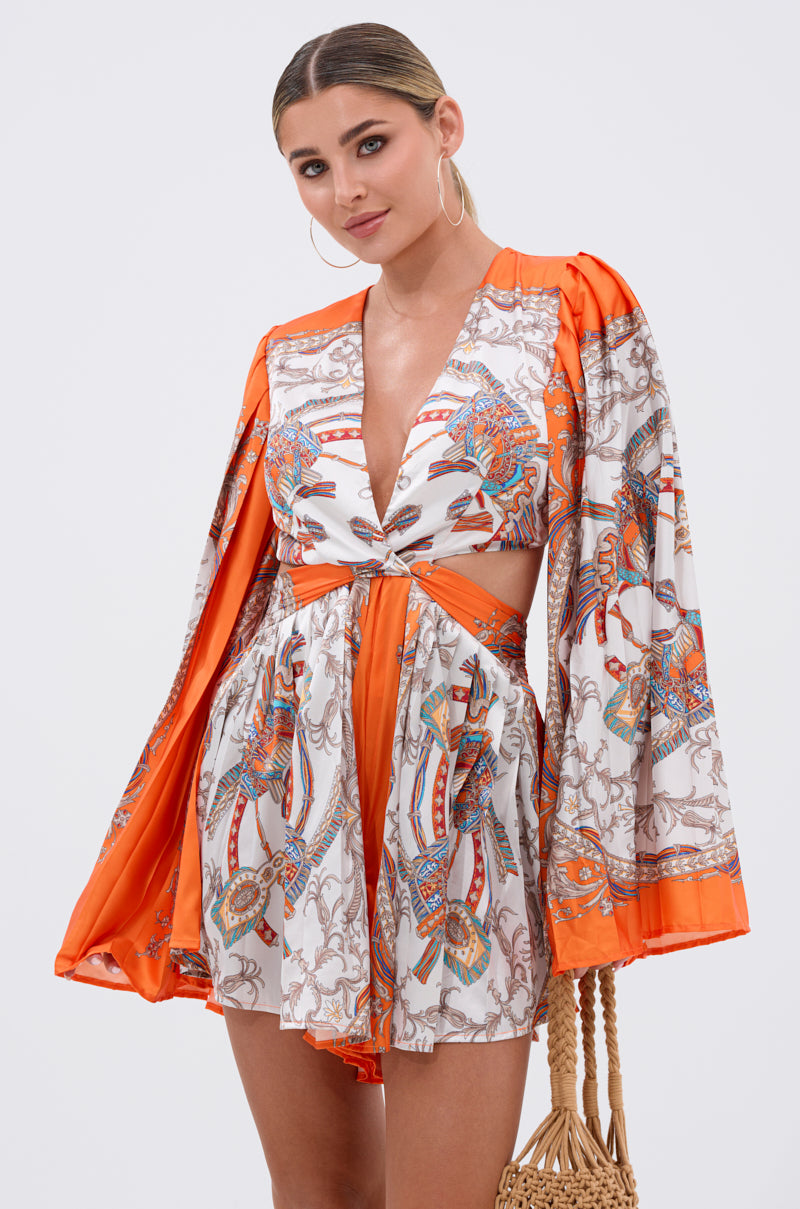 A woman wears the CRAZY IN LOVE SATIN ROMPER, featuring a baroque print, long flowy sleeves, and a deep V-neckline. She holds a woven handbag while standing against a plain white background.