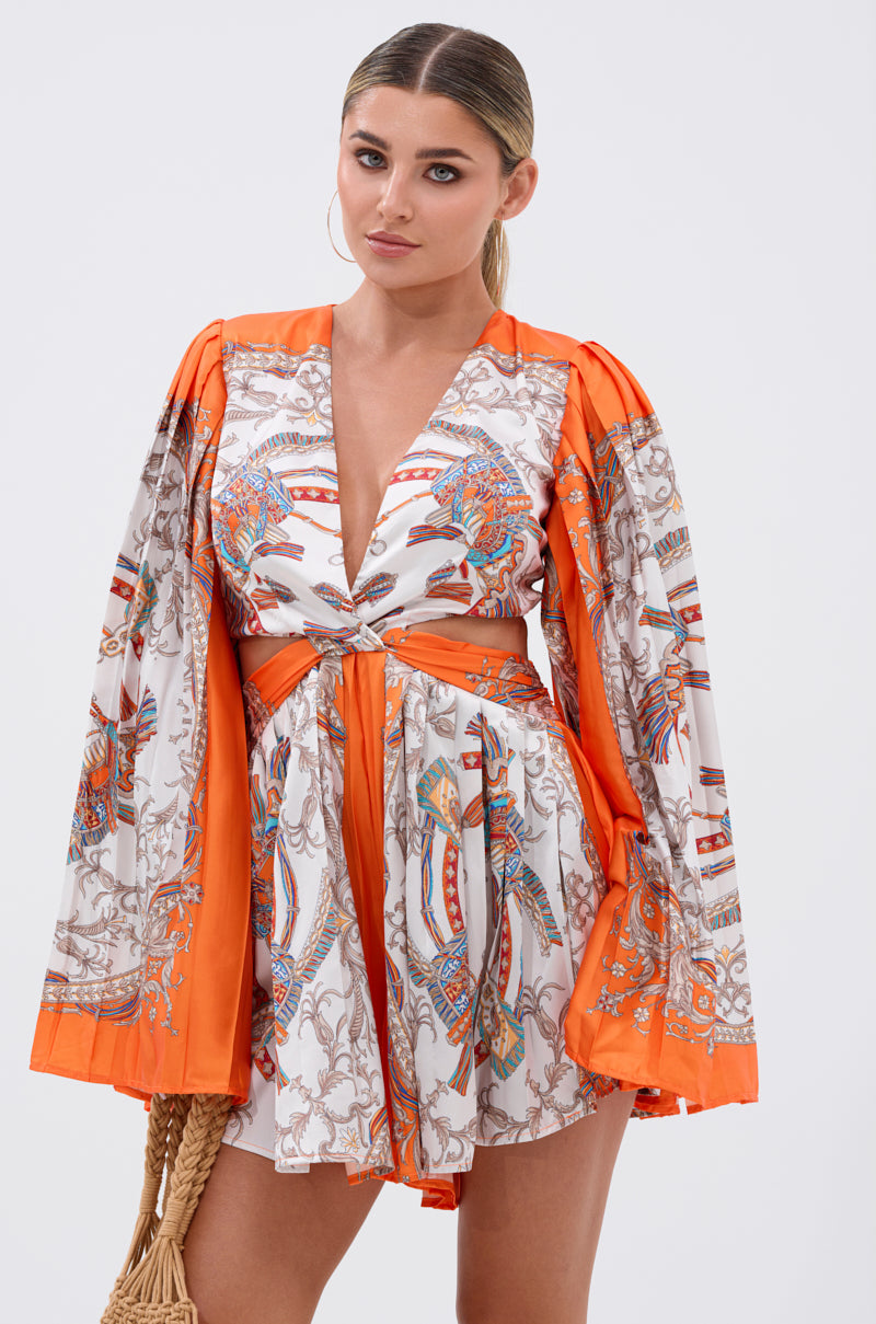 A woman stands against a plain background holding a woven handbag, wearing the CRAZY IN LOVE SATIN ROMPER—a baroque print piece with orange trim, wide sleeves, a deep V-neckline, and a belted waist.