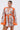 A woman stands against a plain background holding a woven handbag, wearing the CRAZY IN LOVE SATIN ROMPER—a baroque print piece with orange trim, wide sleeves, a deep V-neckline, and a belted waist.