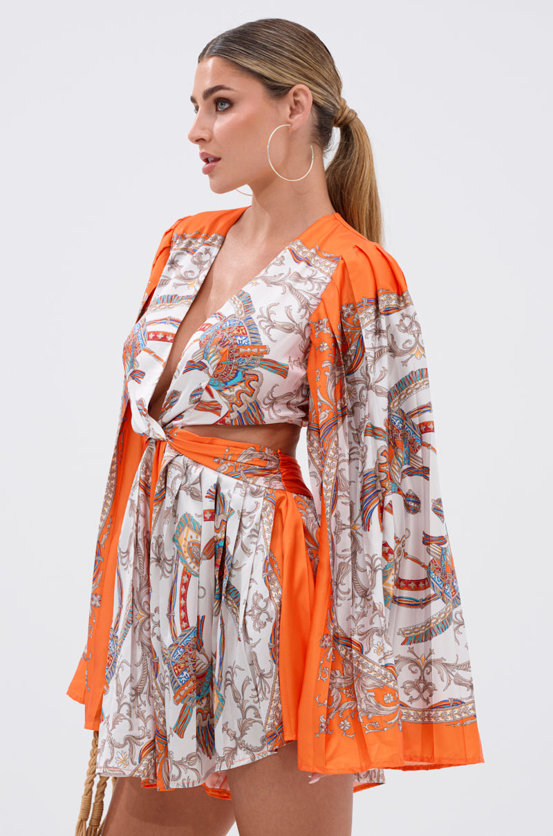 Woman in the CRAZY IN LOVE SATIN ROMPER, a vibrant baroque scarf-print piece with wide orange sleeves, styled with hoop earrings and a low ponytail, posing at an angle against a plain white background.