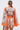 Woman faces away in the CRAZY IN LOVE SATIN ROMPER, featuring a vibrant baroque scarf print and wide sleeves. Her hair is slicked back in a low ponytail, holding a woven bag against a plain light background for a flowy silhouette.