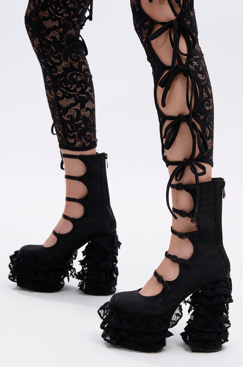 A person wears black lace-up leggings with bold cutouts and bows, paired with AZALEA WANG SPREENE FRILL LACE PUMPS IN BLACK featuring lace ruffles, creating a striking gothic chic look against a plain white background.