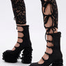 A person wears black lace-up leggings with bold cutouts and bows, paired with AZALEA WANG SPREENE FRILL LACE PUMPS IN BLACK featuring lace ruffles, creating a striking gothic chic look against a plain white background.