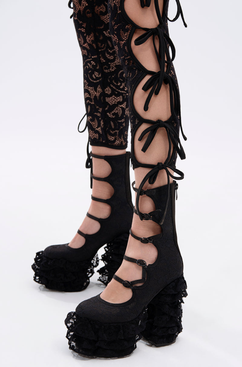 Close-up of a person wearing black lace-up leggings with cutouts and the AZALEA WANG SPREENE FRILL LACE PUMP IN BLACK—gothic platform shoes with chunky heels, multiple straps, and ruffled lace. Set against a plain white background.
