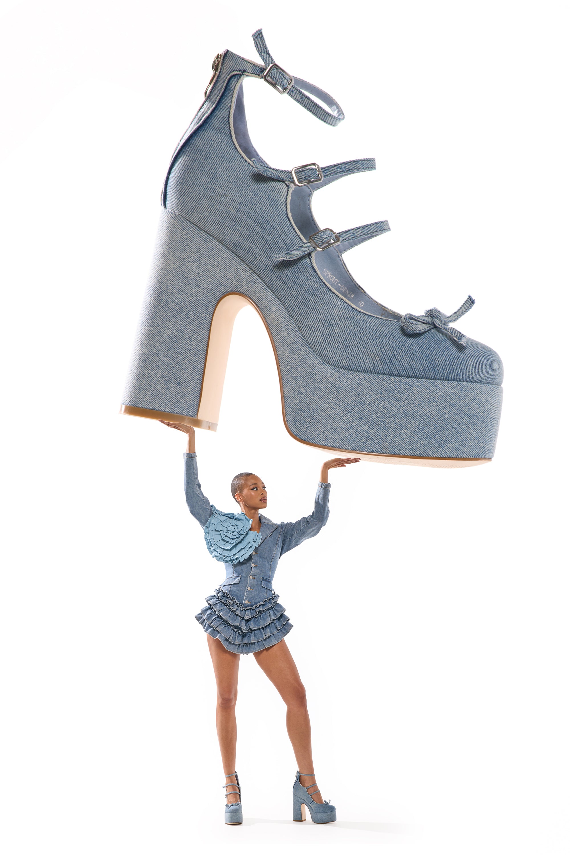 A woman in a ruffled denim outfit and platform heels poses as if lifting the oversized AZALEA WANG SPROUT DENIM CHUNKY PUMP above her head, set against a white background. 0