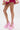 A person wearing a short pink and gold ruffled skirt and AZALEA WANG SPROUT PINK CHUNKY PUMP heels with floral lace and double ankle straps stands against a white background.