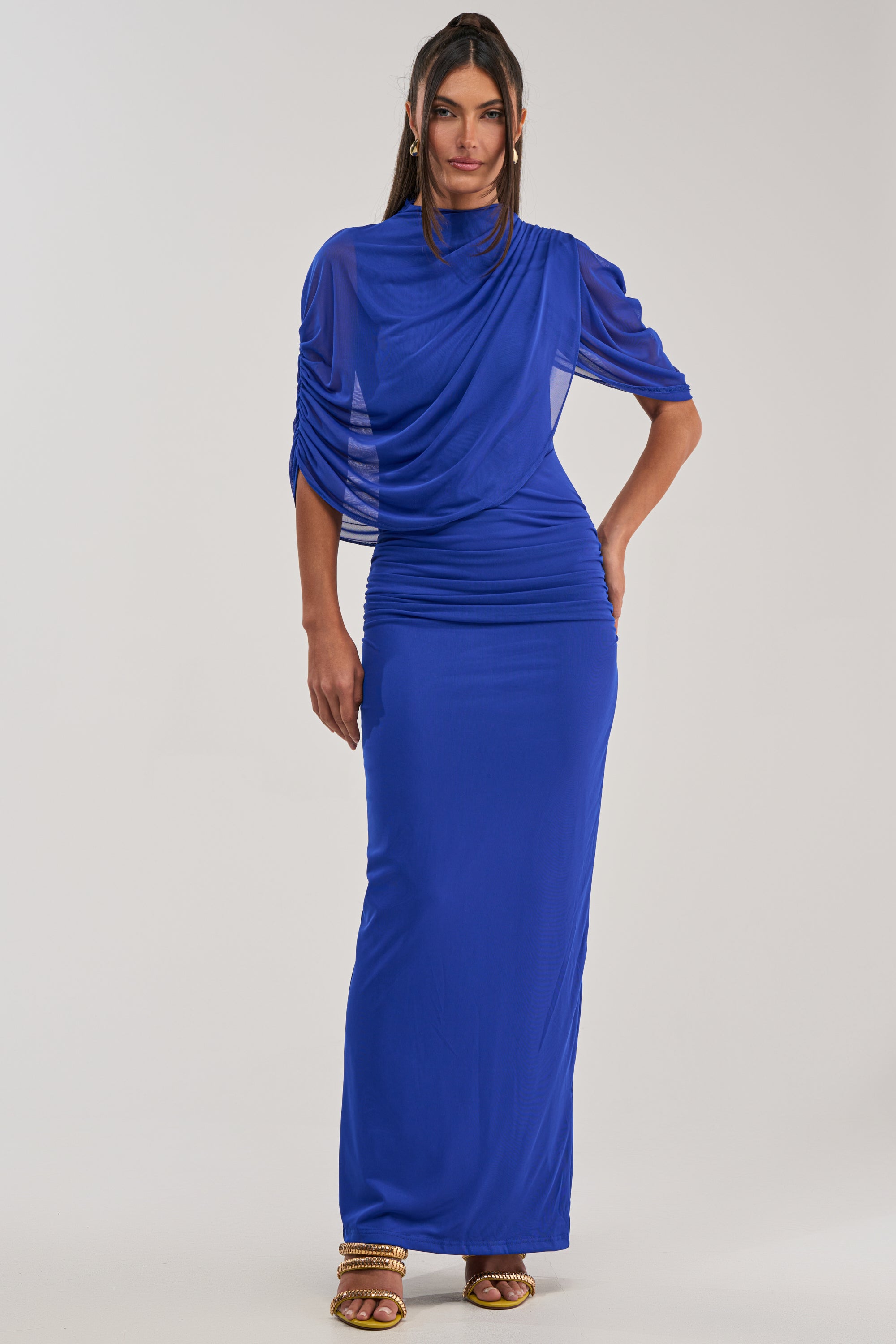 A woman models the GODDESS MENTALITY DRAPED MAXI DRESS in royal blue, featuring ruched detailing and draped sleeves. Styled with long dark hair and gold strappy heels, it’s ideal for formal occasions or wedding guests.