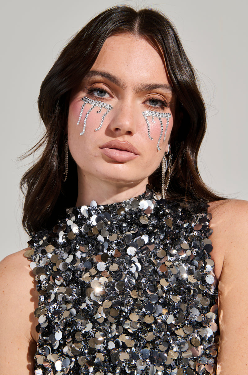 A woman with long brown hair wears a sleeveless, sequined top. She adorns her cheeks with the decorative Tears of Bling Eye Gems, featuring silver and black rhinestones. Hoop earrings complement her neutral expression against a plain gray background.