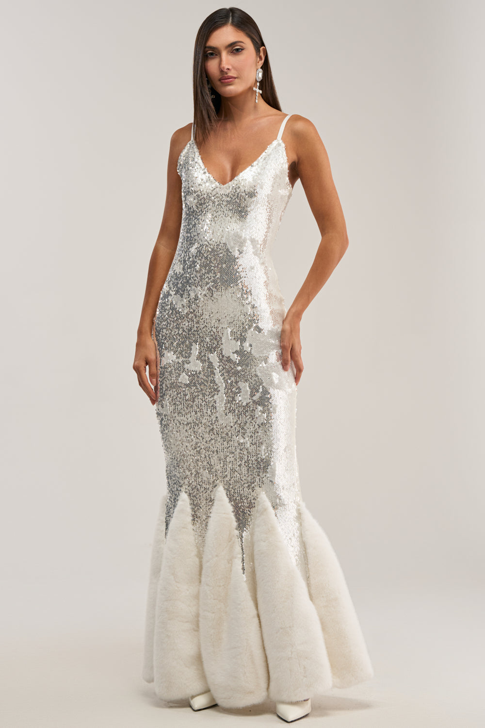 Perfect for holiday parties or formal events, she dazzles in the WORTH THE STARES EMBELLISHED MAXI DRESS—a sleeveless silver sequin gown with a deep V-neck and dramatic white fur-trimmed mermaid hem, styled with earrings and white pointed shoes.