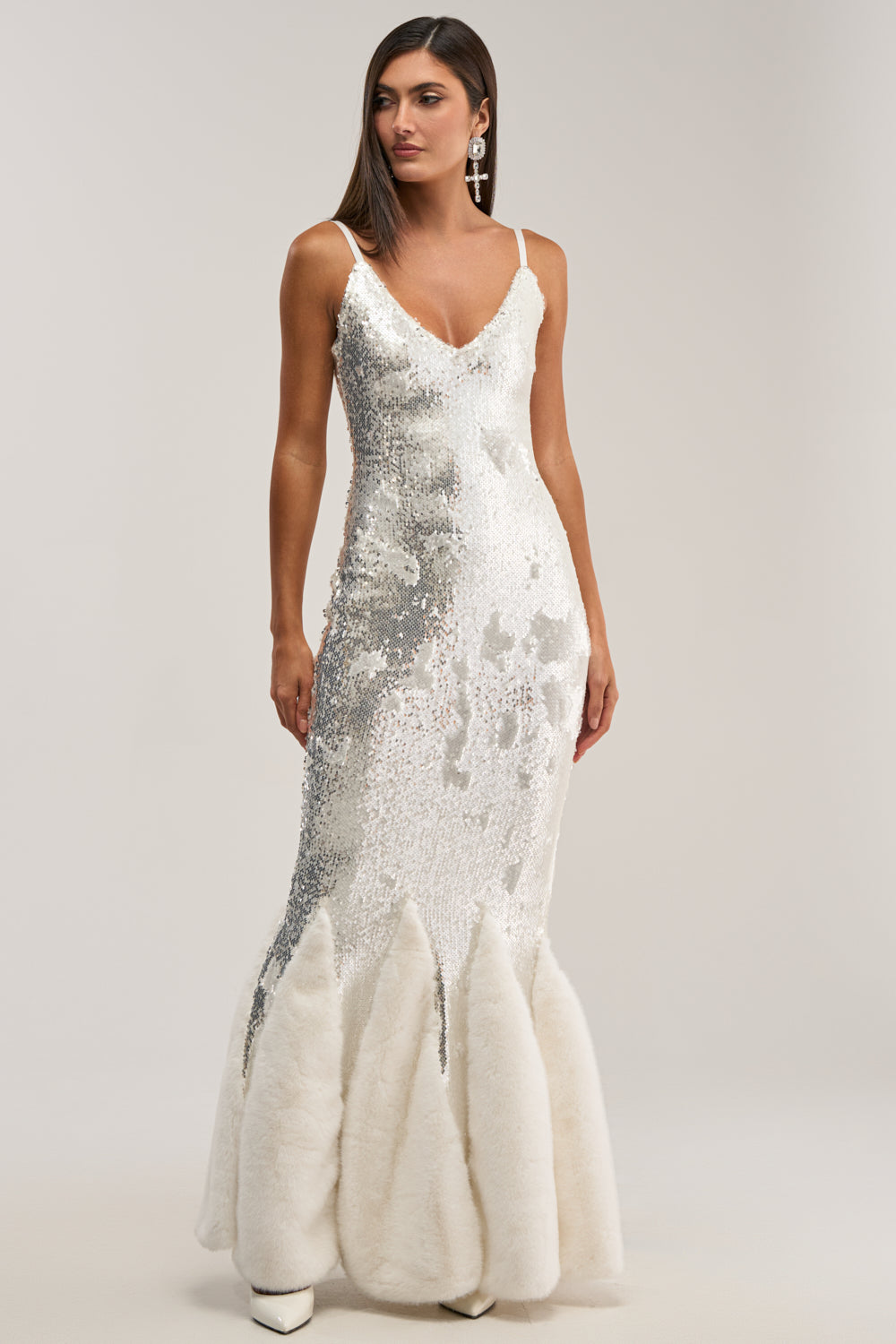 A woman models the WORTH THE STARES EMBELLISHED MAXI DRESS—a sleeveless silver sequin gown with a fitted bodice, flared hem trimmed in white faux fur. Against a plain background, she pairs straight brown hair with a long statement earring for holiday elegance.