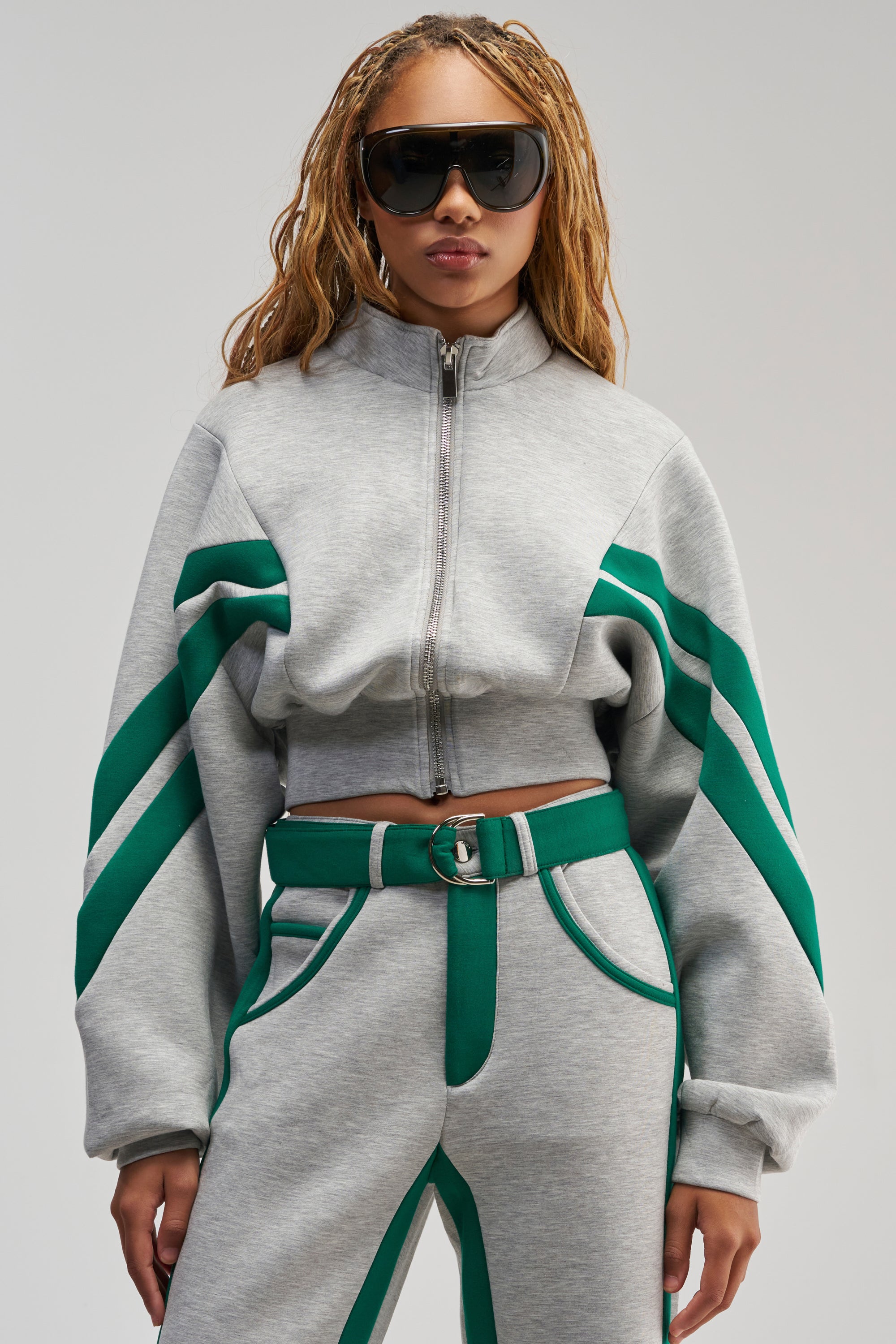 A person styled in the SPORT MODE SCUBA BOMBER IN GREY, featuring green racing stripes and a belt, pairs bold oversized sunglasses with long braided hair and stands confidently against a plain backdrop, epitomizing authentic streetwear.
