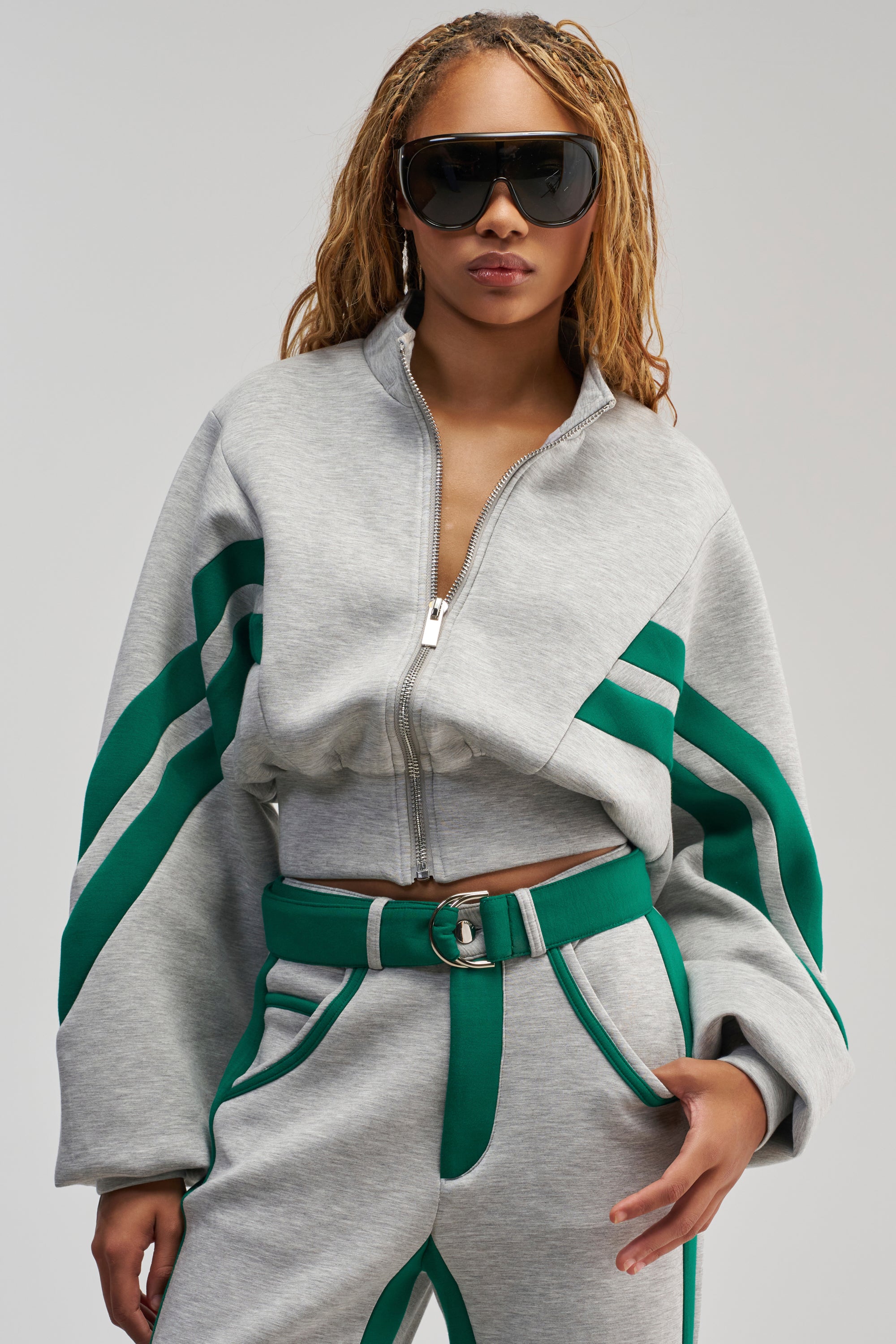 A woman with blond braids wears black sunglasses and the SPORT MODE SCUBA BOMBER IN GREY, featuring bold green racing stripes, a cropped jacket, and matching high-waisted belted pants, posing confidently against a plain background for an effortless streetwear look.