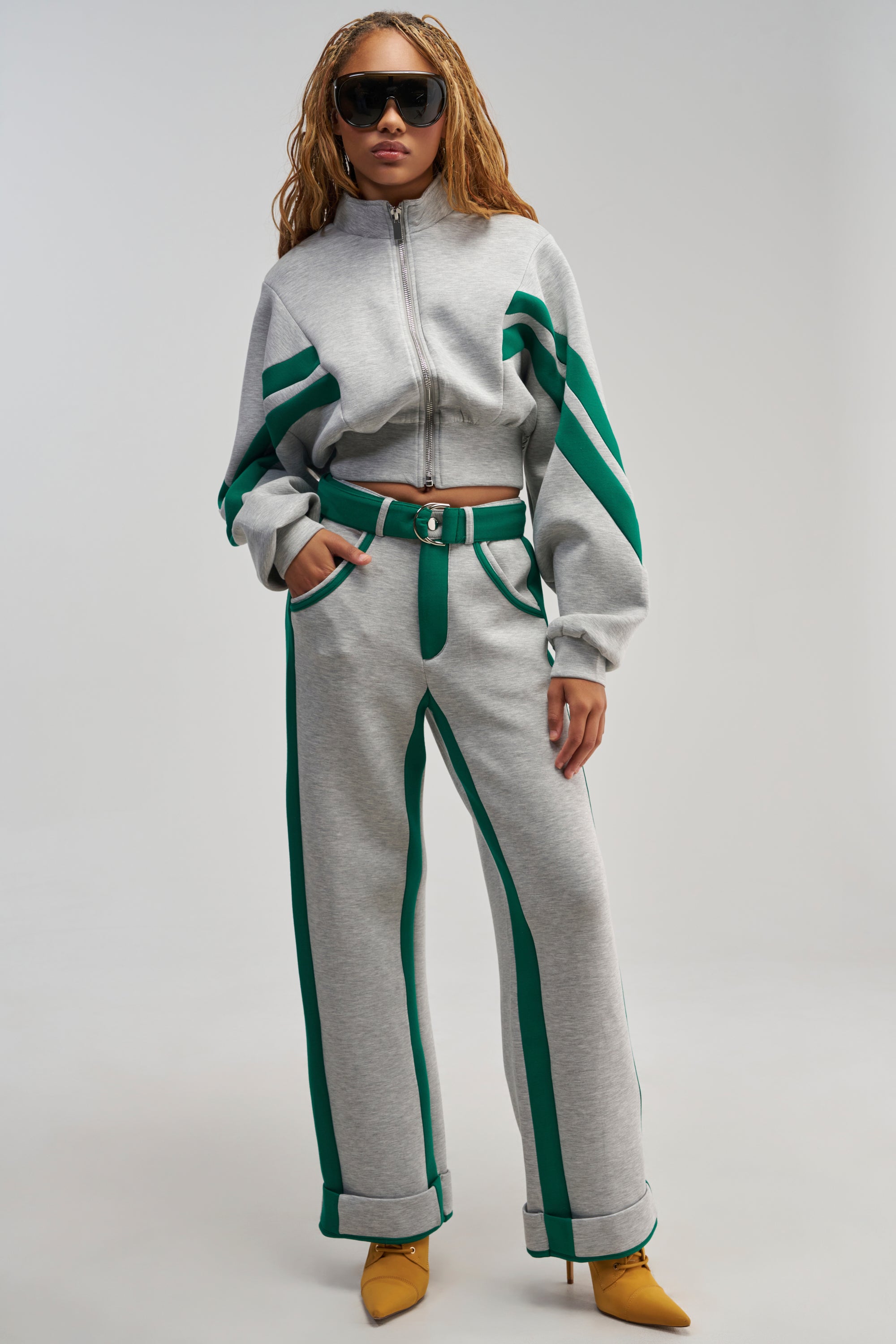 Wearing sunglasses and yellow heels, the person stands against a plain backdrop, dressed in the SPORT MODE SCUBA PANT IN GREY—a pair of high-waisted gray trousers with a belt—paired with a cropped zip-up jacket featuring bold green racing stripes.