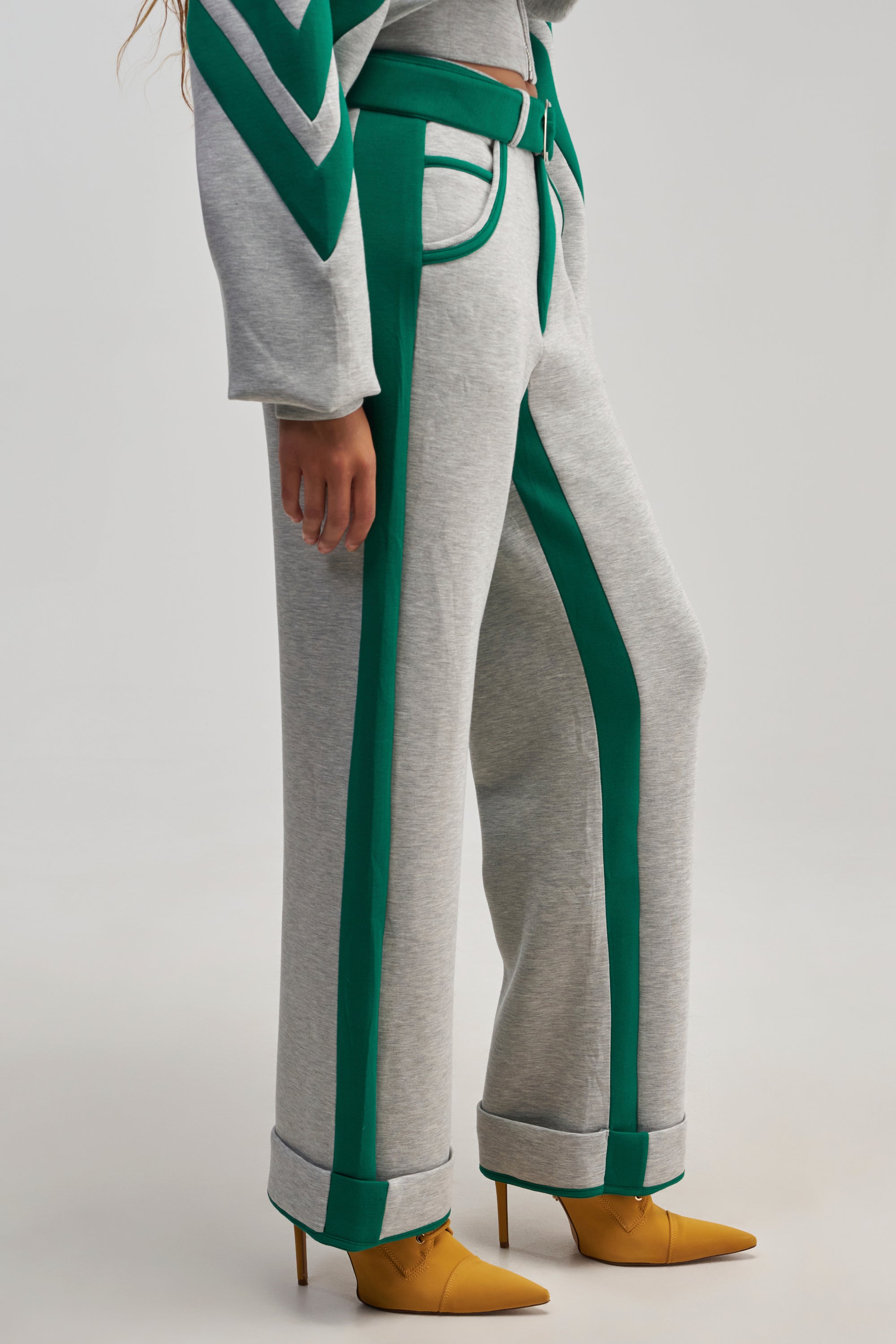 A person exhibits streetwear flair, wearing the SPORT MODE SCUBA PANT IN GREY with green racing stripes, paired with a coordinating top and yellow pointed-toe heels. The image focuses on the lower half of the body against a simple light backdrop.