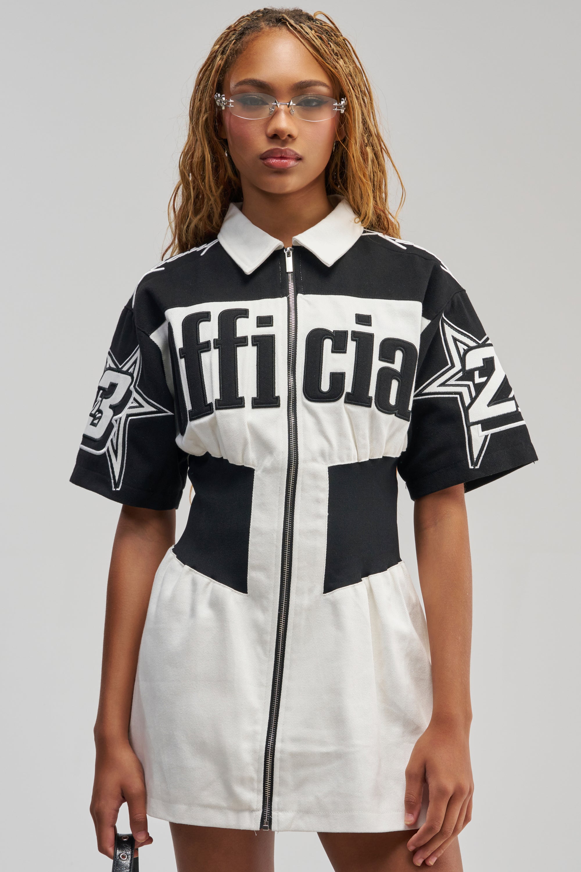 A woman stands against a plain gray background wearing the OFFICIALLY YOURS COLORBLOCK MINI DRESS—a sporty-chic black and white zip-up dress with bold text and star graphics on the sleeves. She accessorizes with clear glasses and holds a small object.