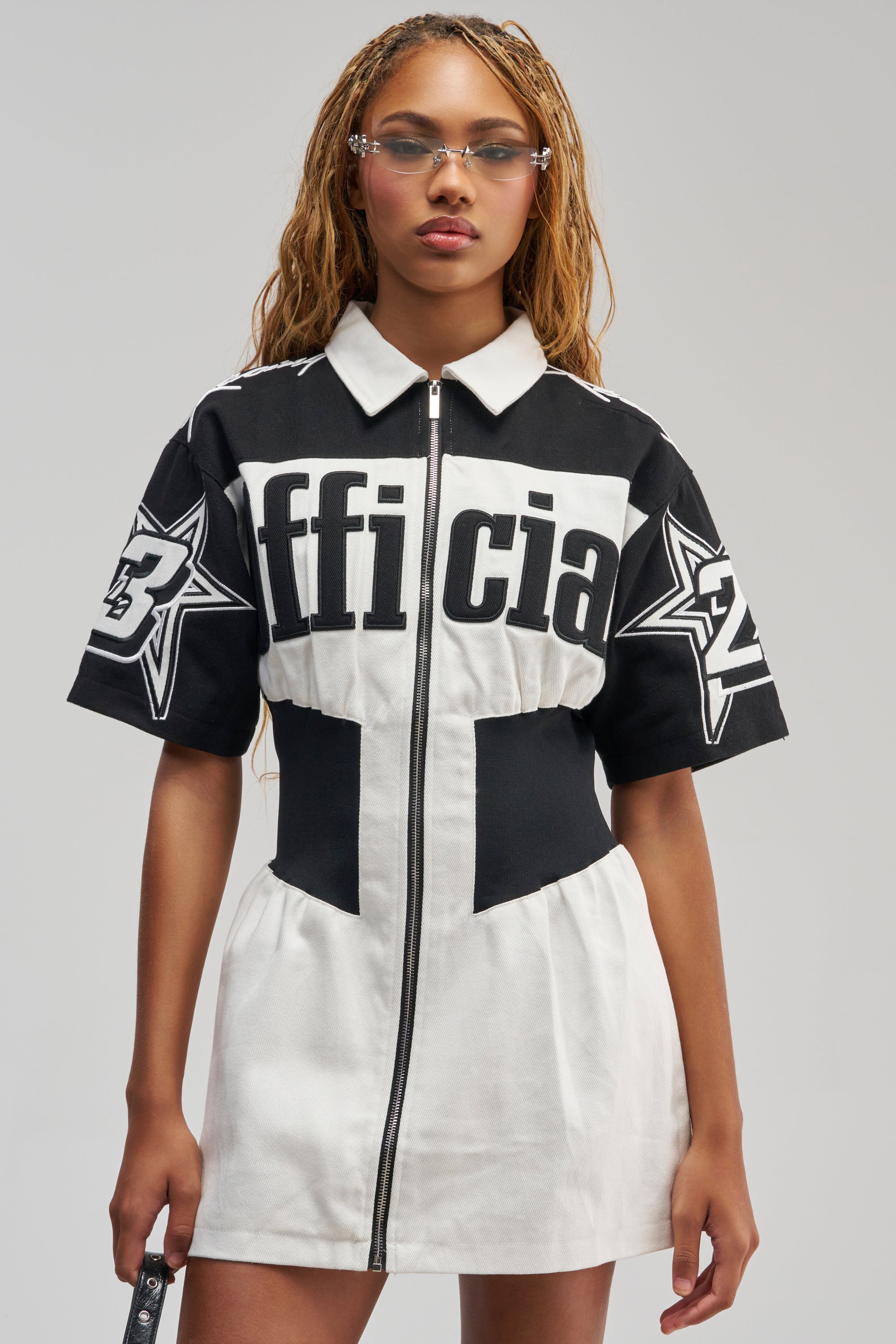 Wearing the OFFICIALLY YOURS COLORBLOCK MINI DRESS, a woman stands against a plain background, holding a phone in her right hand and sporting clear glasses. The sporty-chic mini dress features black-and-white colorblocking, star graphics, and the word “official” across the chest.