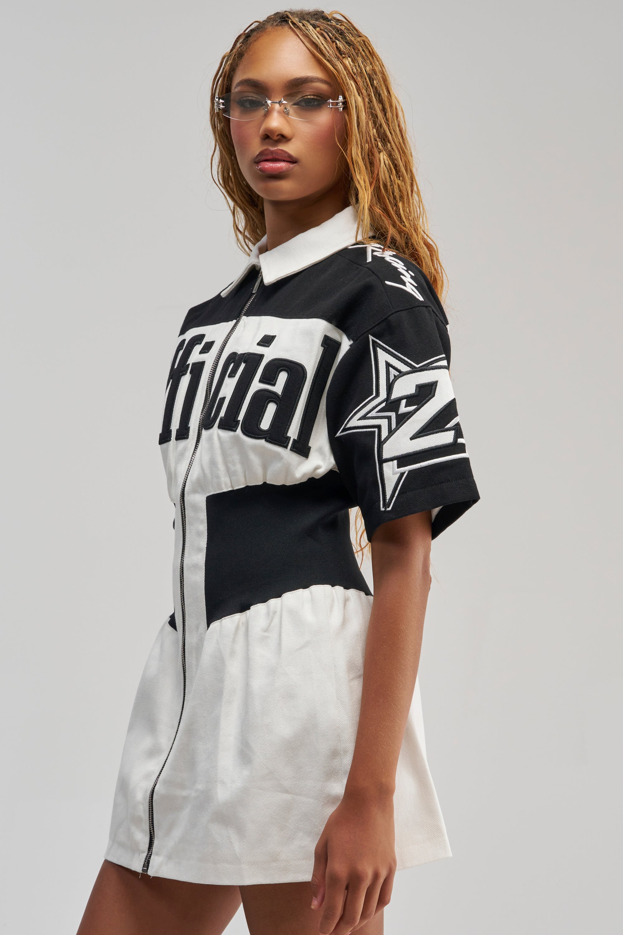 A woman with long blonde braids wears the OFFICIALLY YOURS COLORBLOCK MINI DRESS, a sporty-chic black and white shirt dress featuring bold graphics—including the word “official” and a large star—paired with clear glasses as she stands against a plain background.
